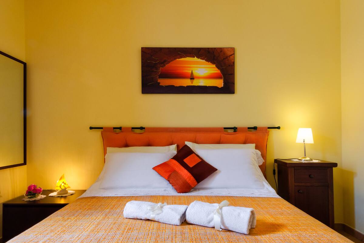 A hotel room with a bed, a painting of a sunset, a nightstand with a lamp, and a neatly made bed with towels.