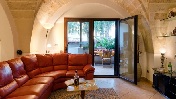 A living room with a leather sofa, a small coffee table with a bottle and glasses, a lamp, and a glass door leading to an outdoor area with a table and chairs. - B&B L'Antica Rimessa (Lecce)