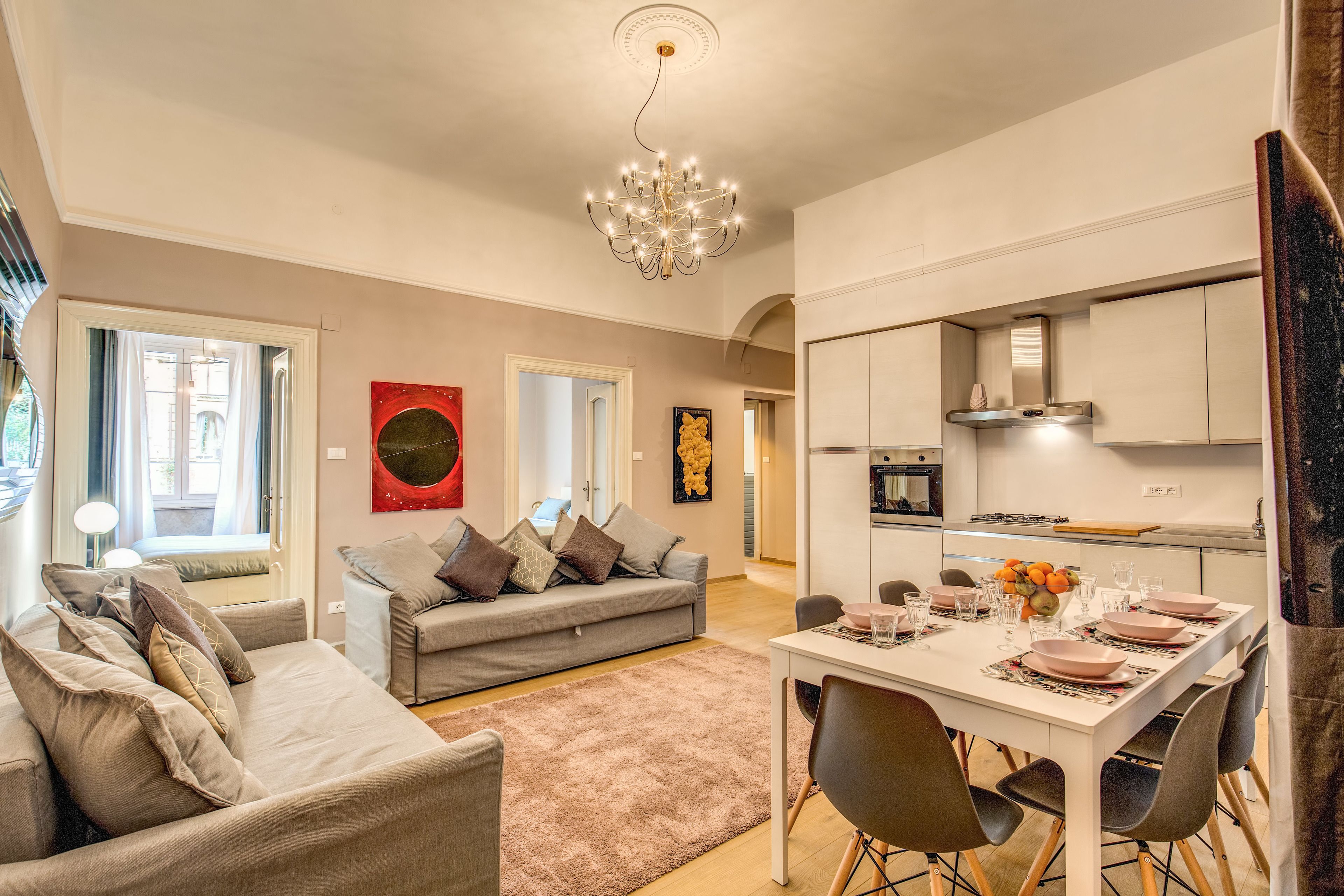 A modern living room with a grey sofa, a dining table set for four, and a kitchen area with white cabinets and a stainless steel range hood.