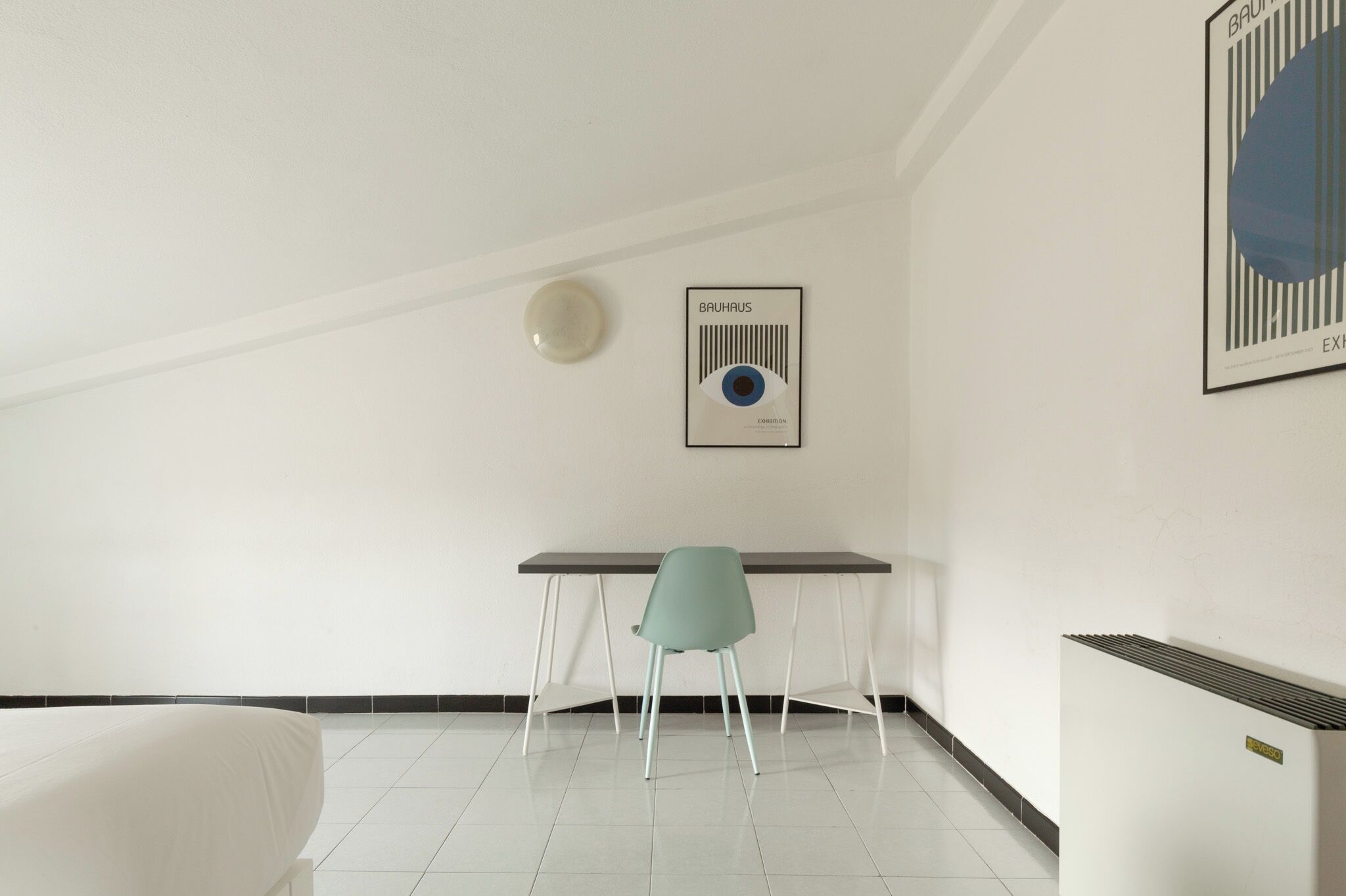 A minimalist room with a bed, a desk, and a chair. There is a framed picture on the wall and a radiator.
