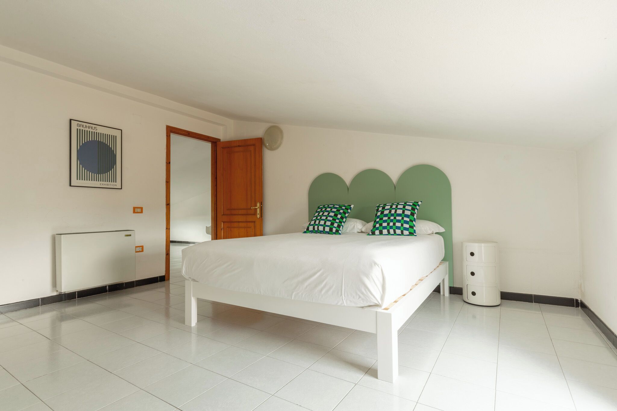 A bedroom with a white bed, green and white checkered pillows, a white bedside table, and a small white trash bin.