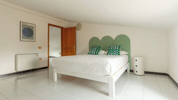 A bedroom with a white bed, green and white checkered pillows, a white bedside table, and a small white trash bin.