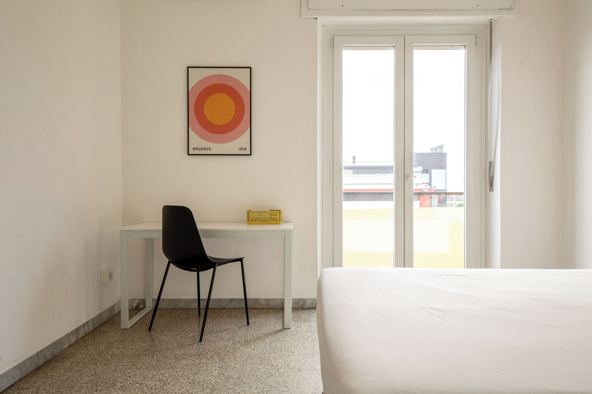 A hotel room with a bed, a desk with a chair, a framed artwork on the wall, and a sliding glass door leading to an outdoor area.