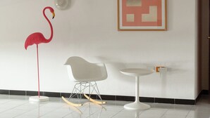 A minimalist room with a white chair, a small round table, a red flamingo decoration, and a framed abstract artwork on the wall. - Spacebility Airport-  self check-in (Cagliari)