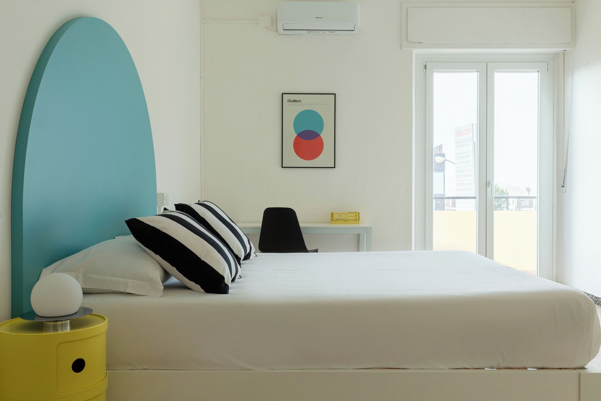 Habitación de hotel con cama, escritorio, silla, ventana grande y aire acondicionado instalado en la pared.