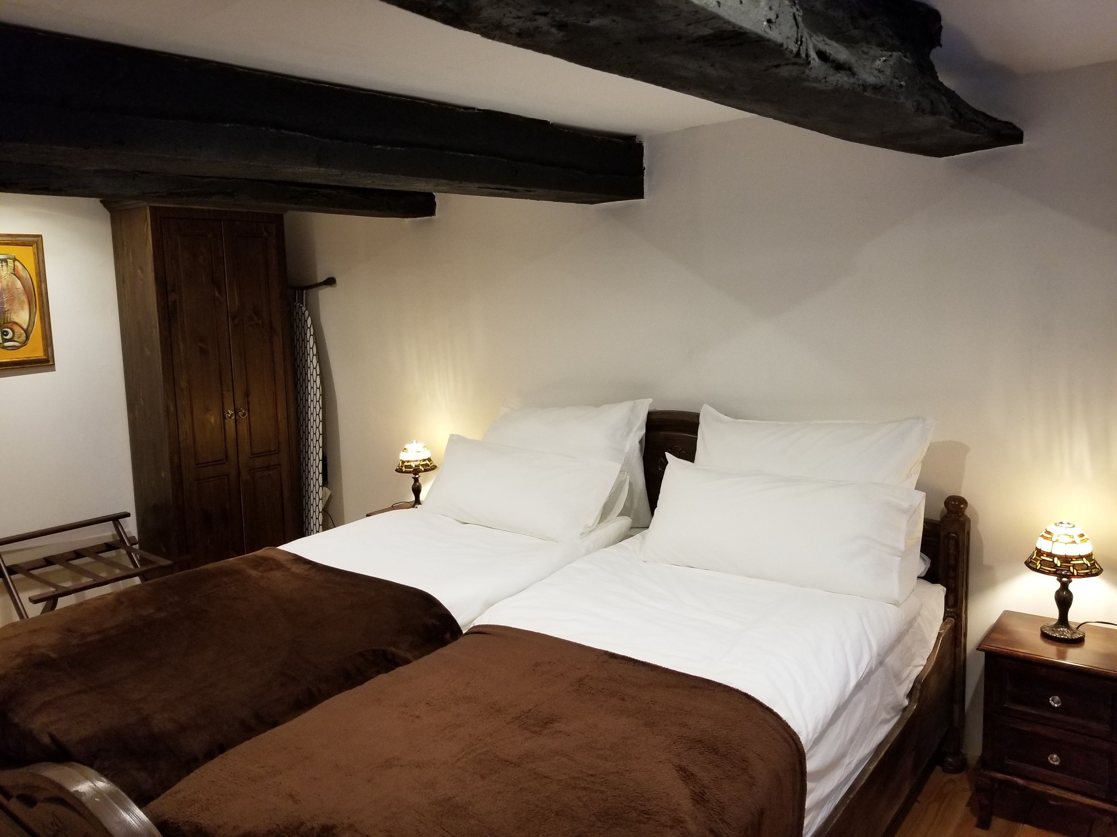 A double bed with white linens and brown throw pillows, two bedside lamps, a wooden headboard, and a dark ceiling beam.