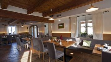 Ein gemütliches Restaurant mit Holzbalken, einem Essbereich mit Tischen und Stühlen sowie einem großen Fenster mit Vorhängen.