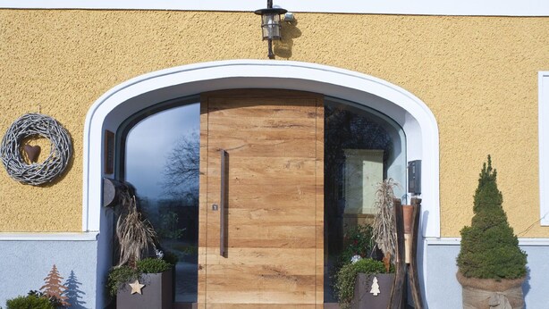 Entrada de un edificio moderno con puerta de madera, una corona a la izquierda y dos macetas a ambos lados.
