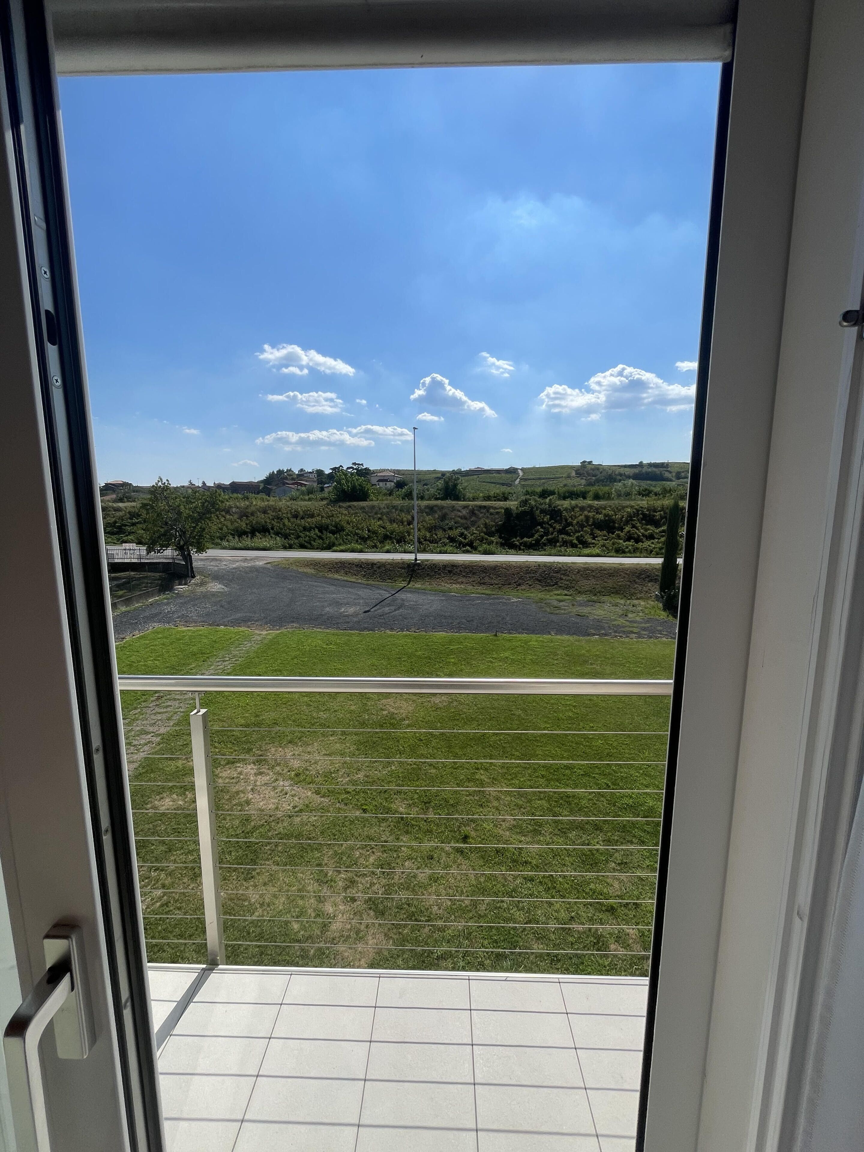 Blick vom Balkon auf einen bewachsenen Bereich und eine Straße in der Ferne, mit blauem Himmel.