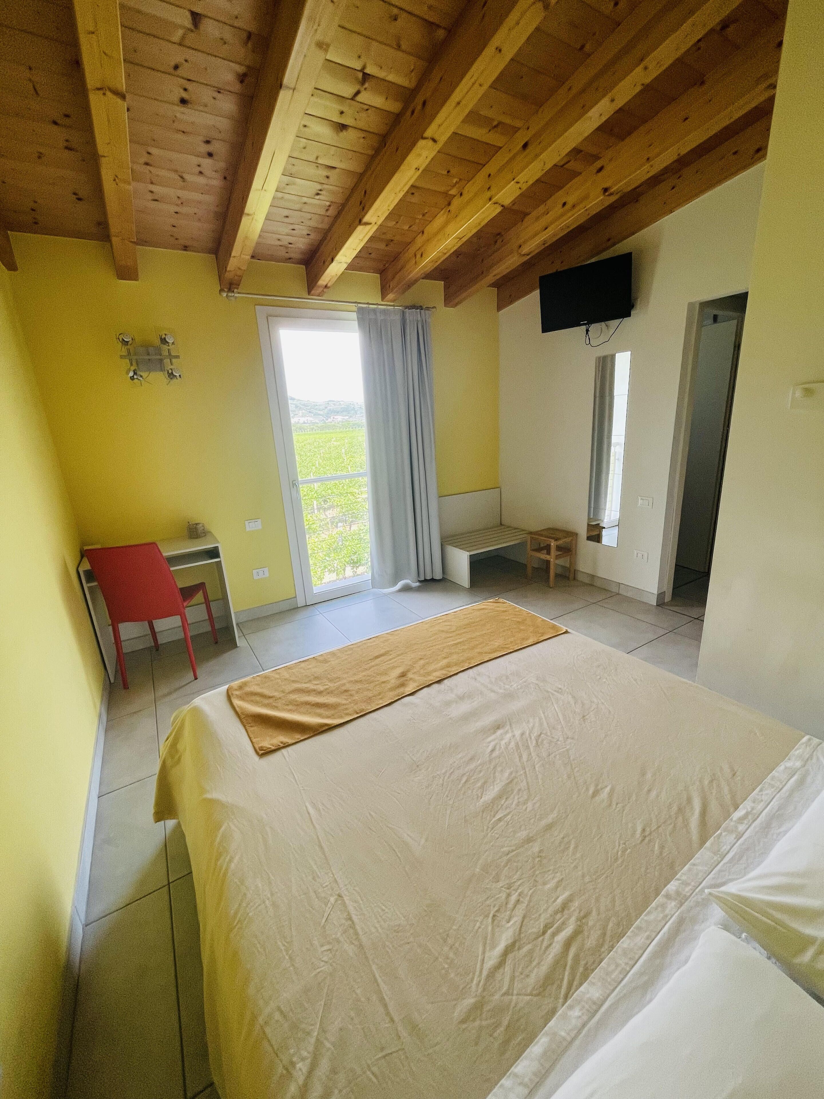 A bedroom with a bed, a desk, and a chair. There is a window with curtains, a TV mounted on the wall, and a door leading to another room.