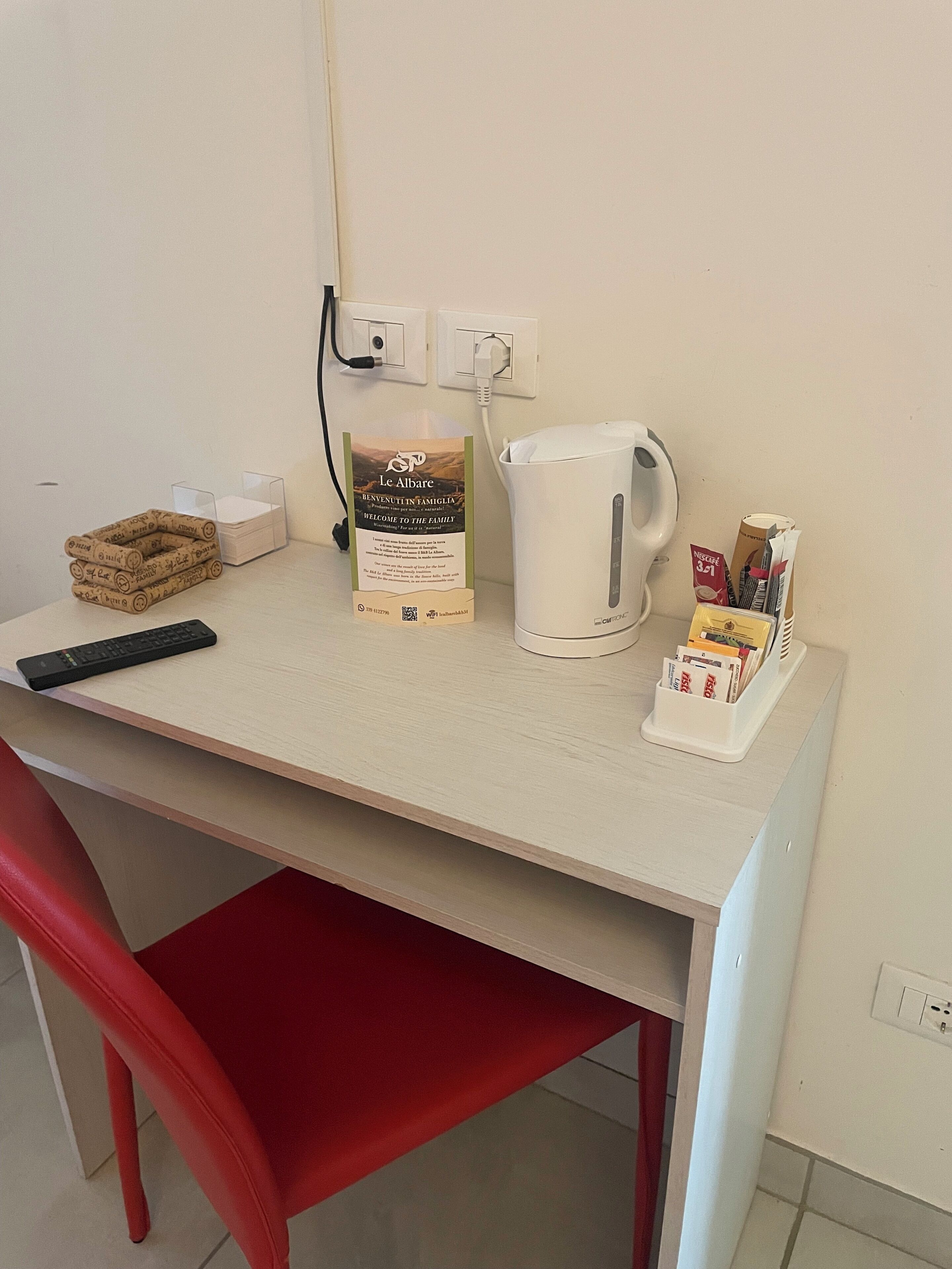 A hotel room with a desk, a red chair, a coffee maker, a kettle, a sign, and a remote control.