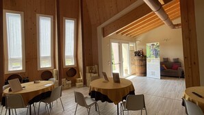 A spacious room with wooden floors and high ceilings, featuring large windows and a central table with chairs.
