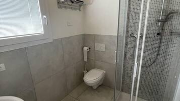 Un baño con inodoro, lavamanos, ducha con puerta de cristal y ventana con persianas.
