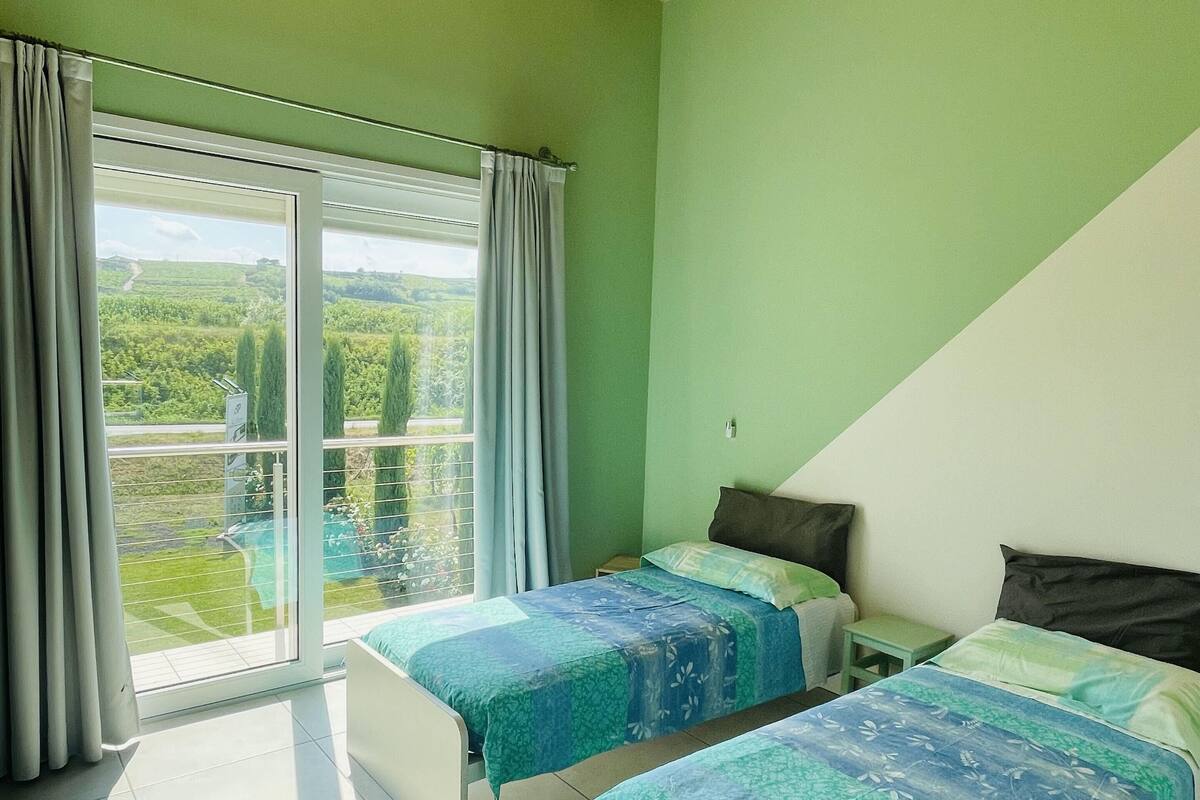 A bedroom with two beds, a large window with curtains, and a view of greenery outside.