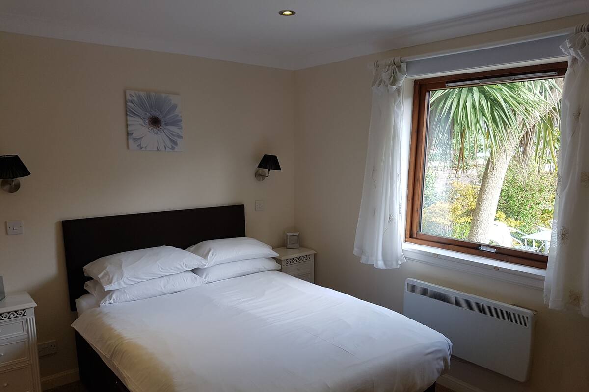 A bedroom with a bed, a nightstand, a wall clock, a window with curtains, and a radiator.