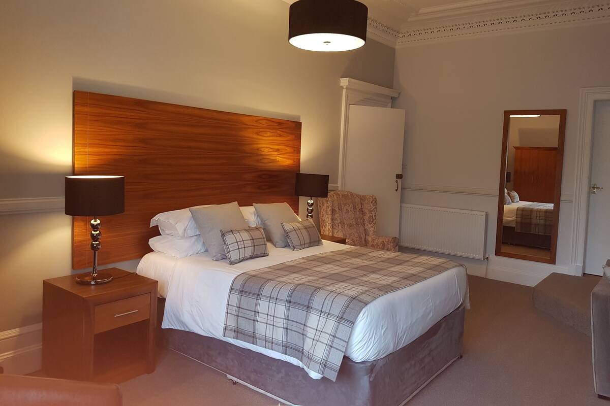 A bedroom with a wooden headboard, a bed with white linens and plaid bedding, two bedside lamps, a nightstand, a mirror, and a radiator.