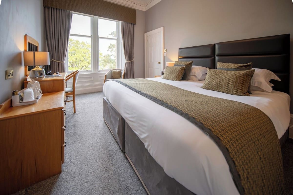 A hotel room with a large bed, a desk with a chair, a window with curtains, and a door.