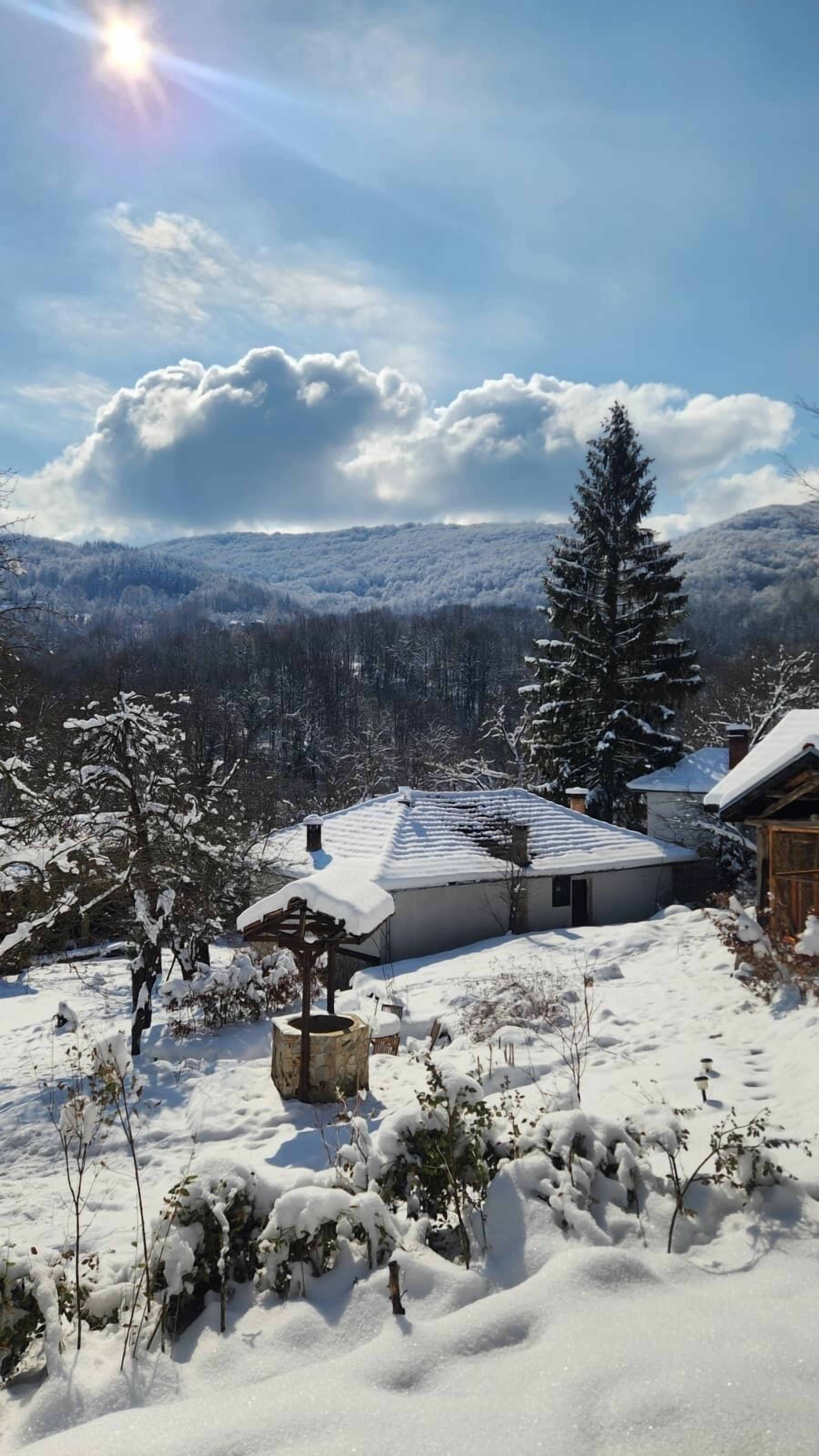 A snow-covered landscape with a house featuring a sloped roof and a stone structure in the foreground.