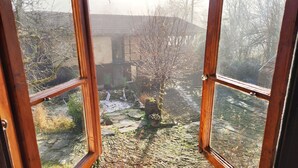 A view through a wooden-framed window of a stone-paved garden with a small tree, a wooden structure, and a stone wall. - Little River Guest House (Gabrovo)