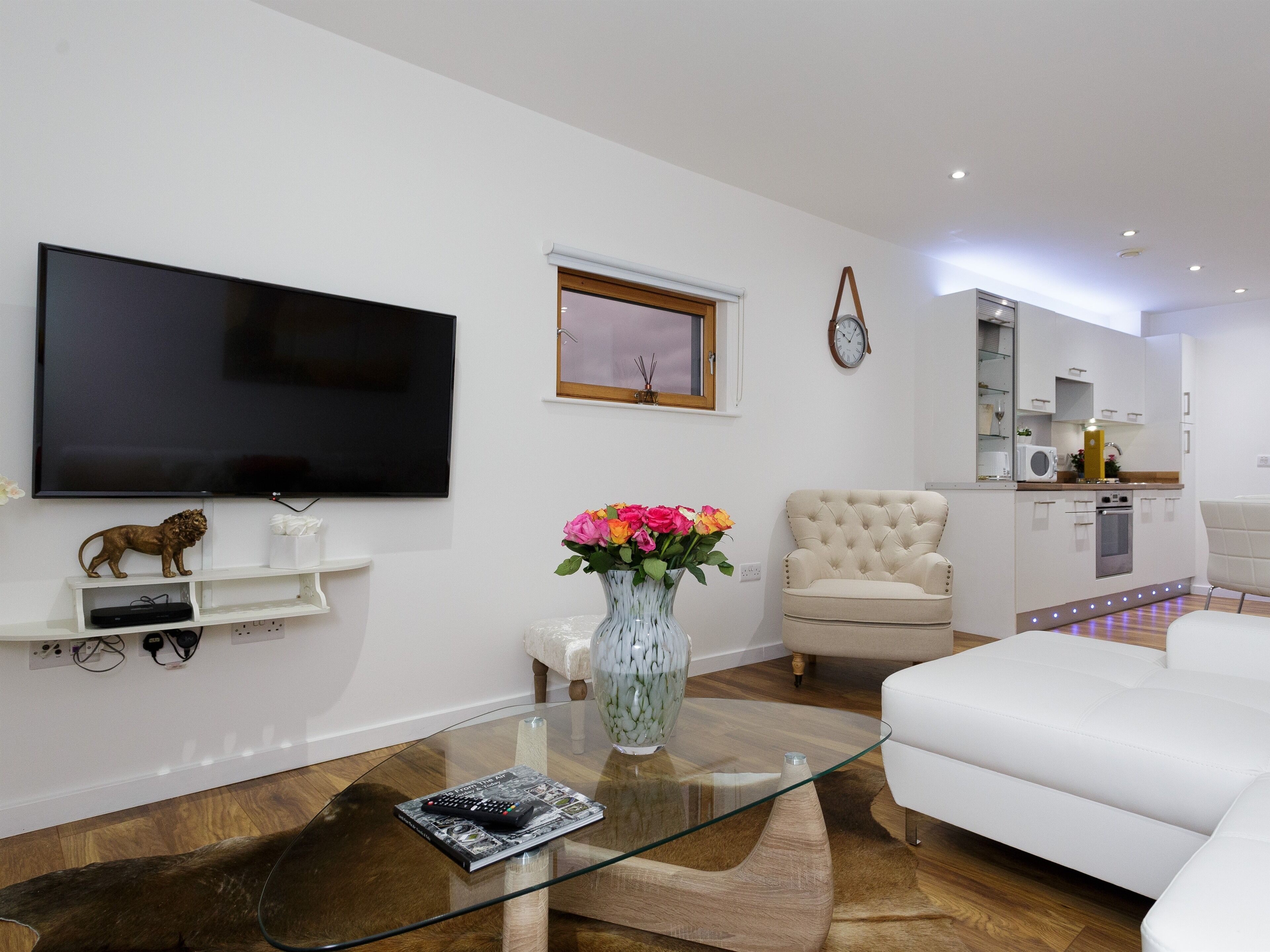 Ein modernes Wohnzimmer mit einem Flachbildfernseher, einem Glastisch, einem weißen Sofa, einem beige farbenen Sessel und einer kleinen Küchenzeile mit einer Mikrowelle und einer Wanduhr.
