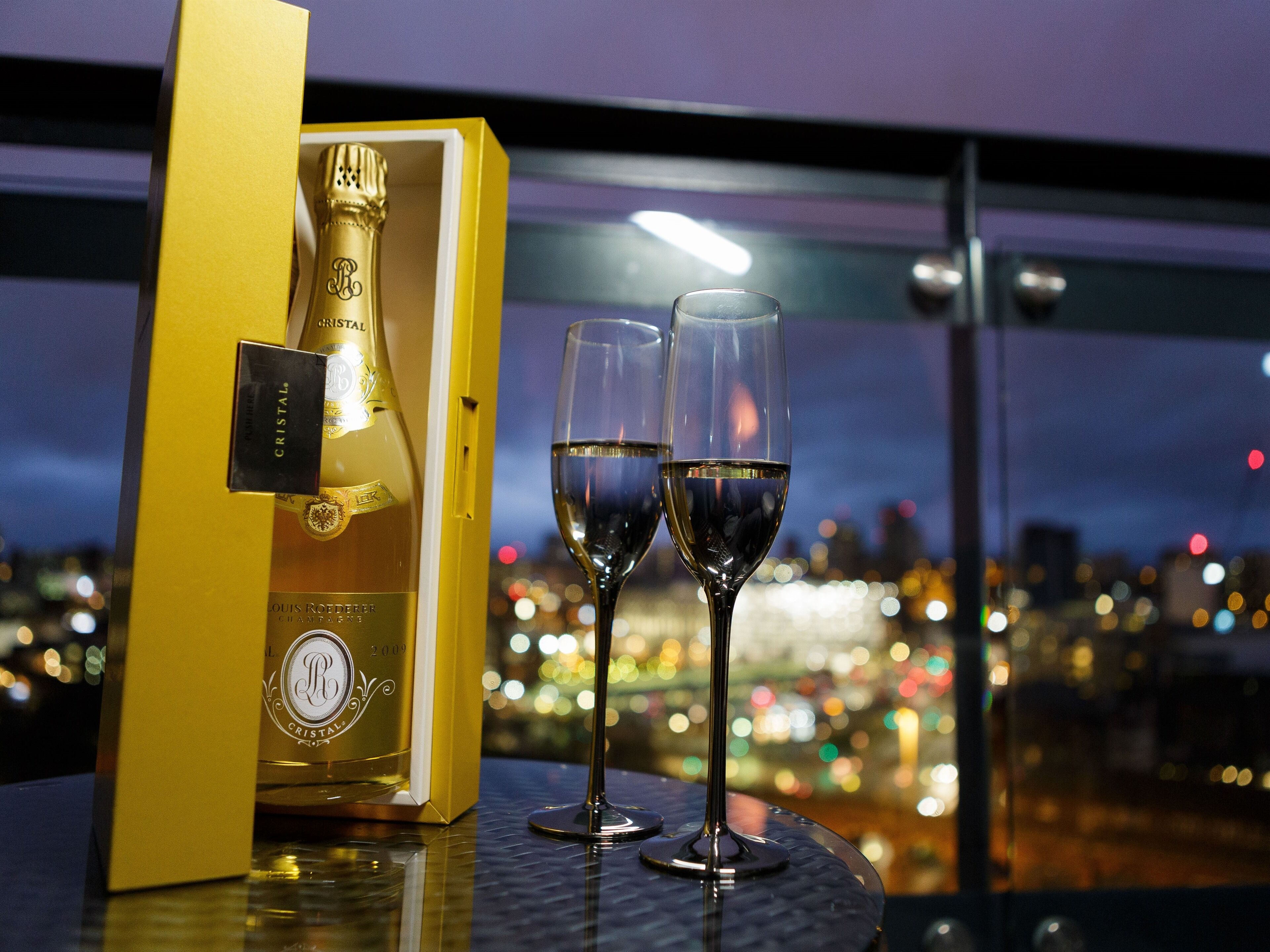 Una botella de champán Cristal en una caja amarilla, dos copas de champán y una vista panorámica de la ciudad a través de una ventana.