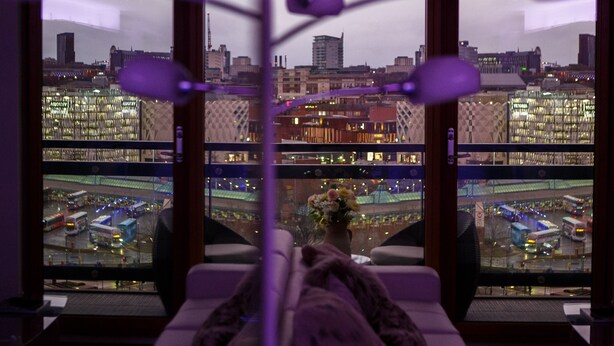 A room with a view of a cityscape, featuring modern buildings and a busy street with buses. - Live in Leeds Sky Penthouse (Leeds)