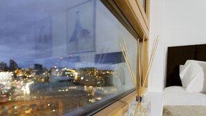 A bedroom with a view of a cityscape at night, a bedside table with a reed diffuser, and a bed with a white cover. - Live in Leeds Sky Penthouse (Leeds)