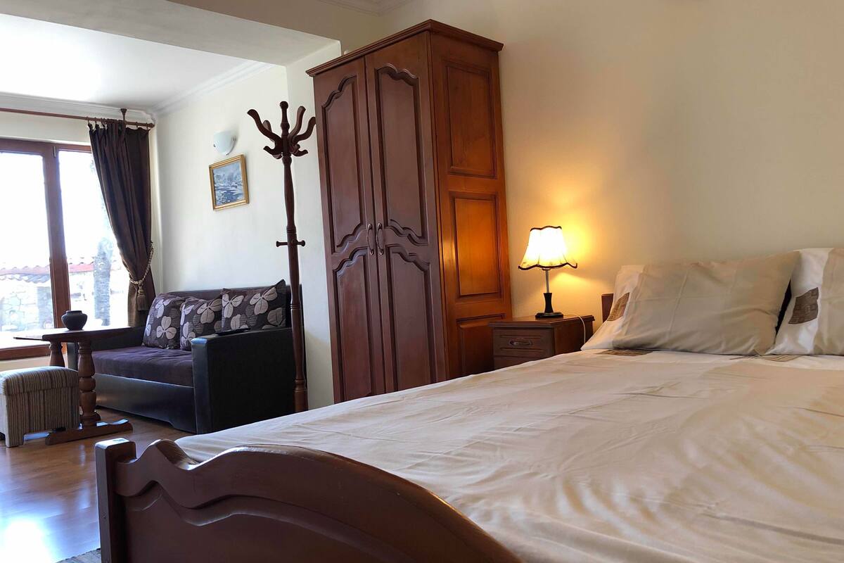 A hotel room with a bed, a wooden wardrobe, a sofa, a nightstand with a lamp, and a window with curtains.