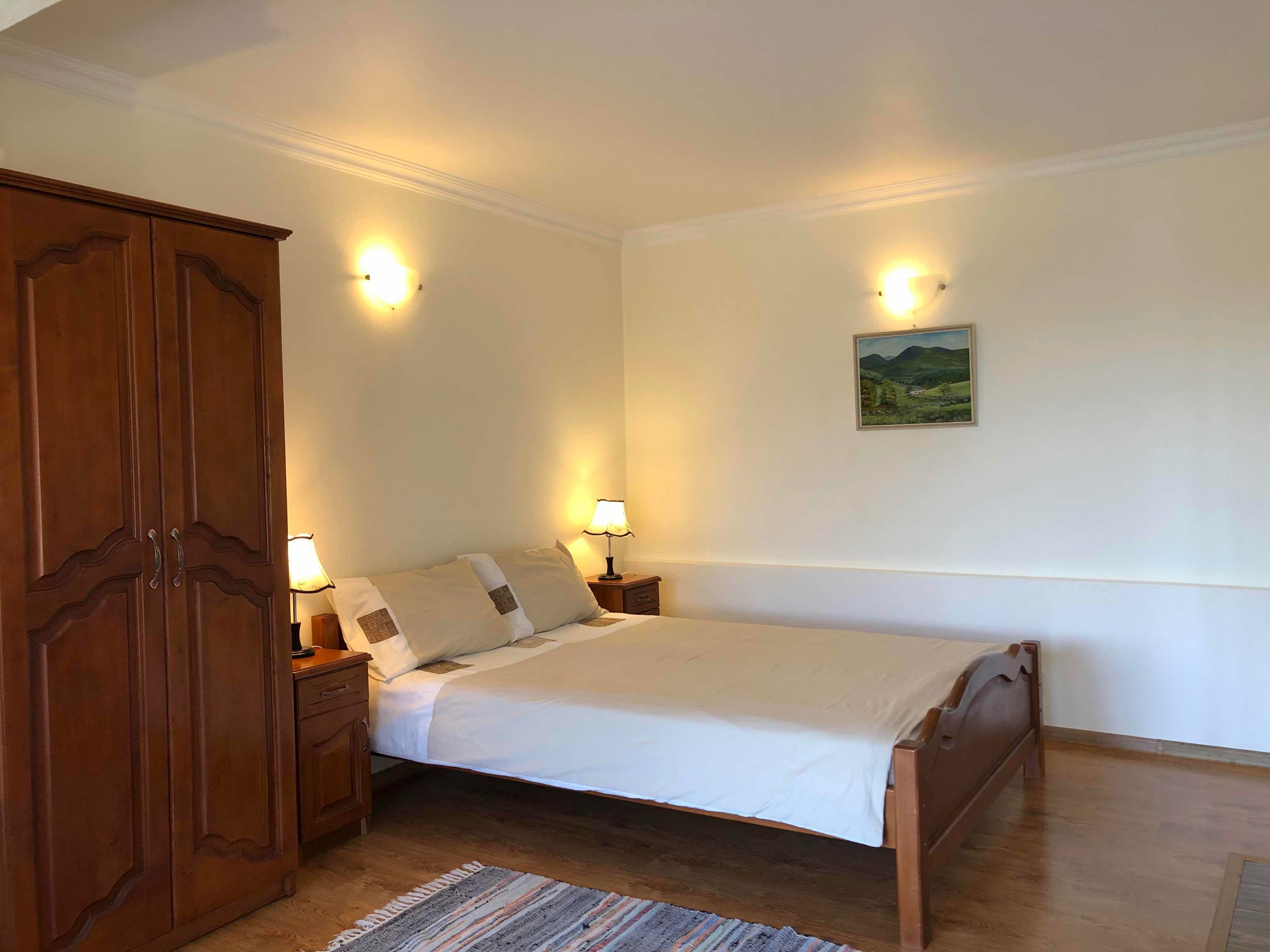 Une chambre à coucher avec un lit en bois, deux tables de chevet avec des lampes, une armoire en bois et un tableau représentant un paysage accroché au mur.
