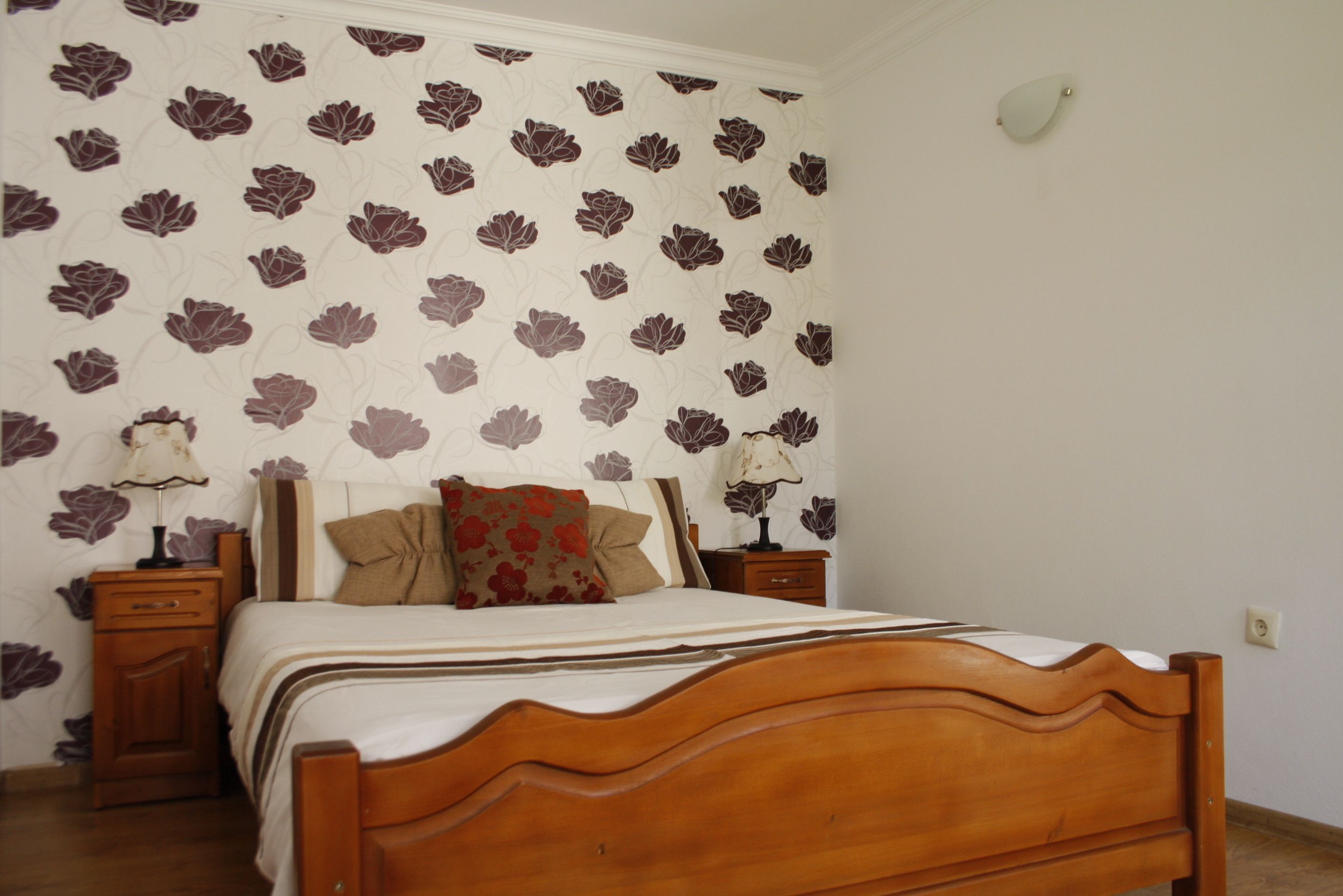 Un dormitorio con una cama de madera, papel tapiz floral, dos mesitas de noche con lámparas y una mesita auxiliar.