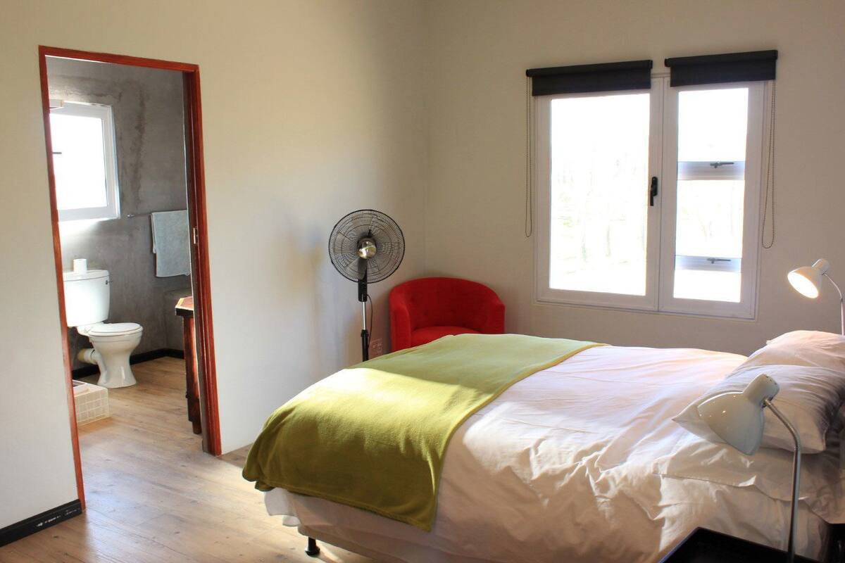 A bedroom with a bed, a red chair, a standing fan, a window with blinds, and a bathroom visible through an open door.