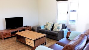 A living room with a wooden coffee table, a grey sofa with patterned cushions, a flat-screen TV on a wooden stand, and a window with blinds. - Village Lodge Paarl (Paarl)