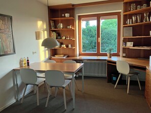 A modern kitchen with a dining area, wooden shelves with kitchenware, a radiator, and a window with a view of greenery.