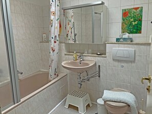 A bathroom with a bathtub, a sink, a toilet, a shower curtain, a mirror, a small stool, and a painting on the wall.