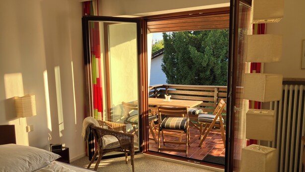 A hotel room with a bed, a chair, a table, and a sliding glass door leading to an outdoor seating area. - Absolutely quiet 3 Room Apartment "At Isar River Park",  near Munichs Centre (Grünwald)