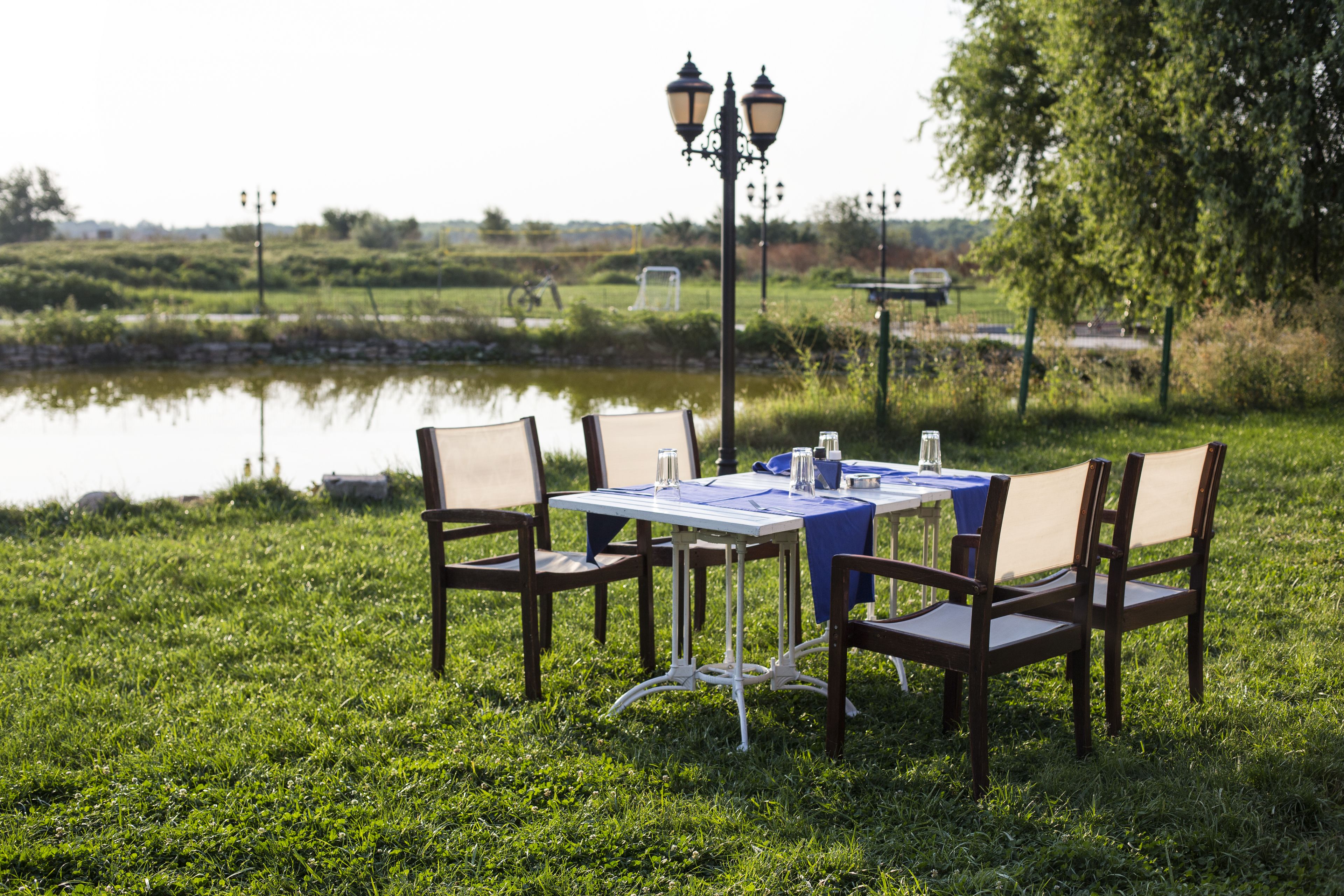 An outdoor dining area with a table set for two, surrounded by chairs, near a body of water and lamp posts.