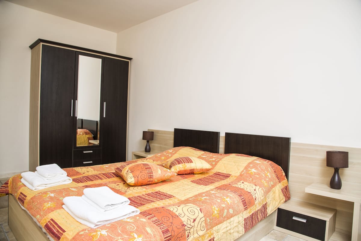 A neatly made bed with a patterned bedspread, a large wardrobe, and a bedside table with a lamp.