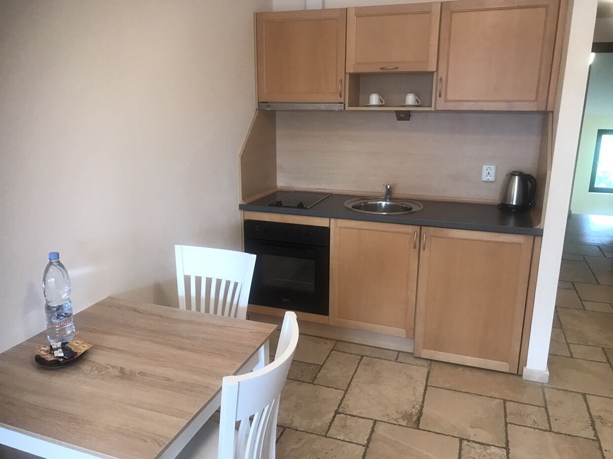 Une cuisine de taille réduite, avec des meubles en bois, une cuisinière noire et un évier. On peut voir une chaise blanche et une table sur laquelle se trouve une bouteille d’eau.