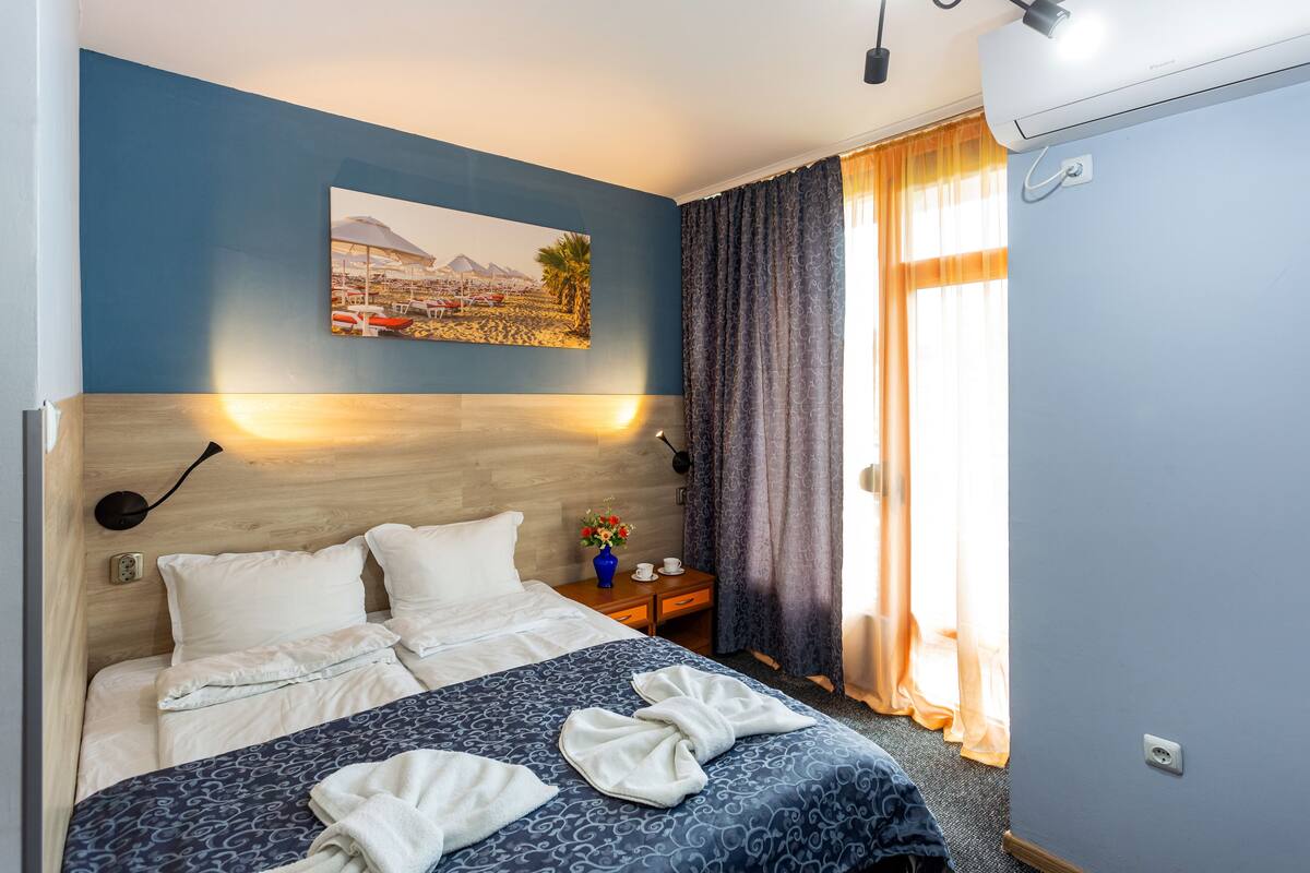 A hotel room with a bed, a nightstand, a vase with flowers, and a painting on the wall.