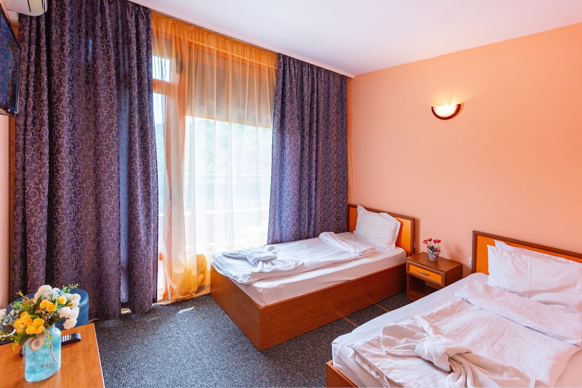 A hotel room with two beds, a desk with a vase of flowers, and a window with curtains.