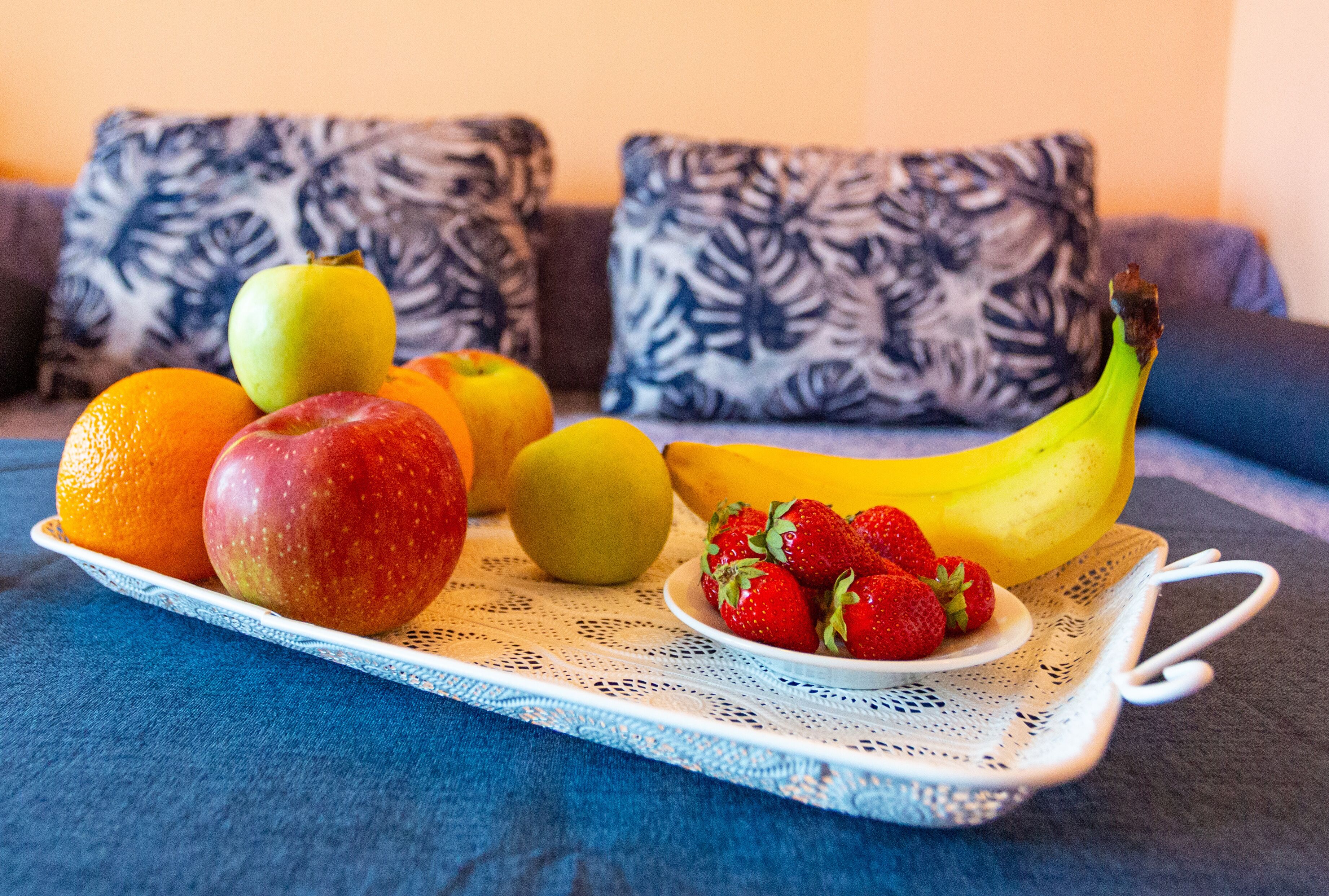 Uma bandeja com frutas, incluindo maçãs, laranjas, uma banana e morangos, sobre um sofá azul com almofadas estampadas.
