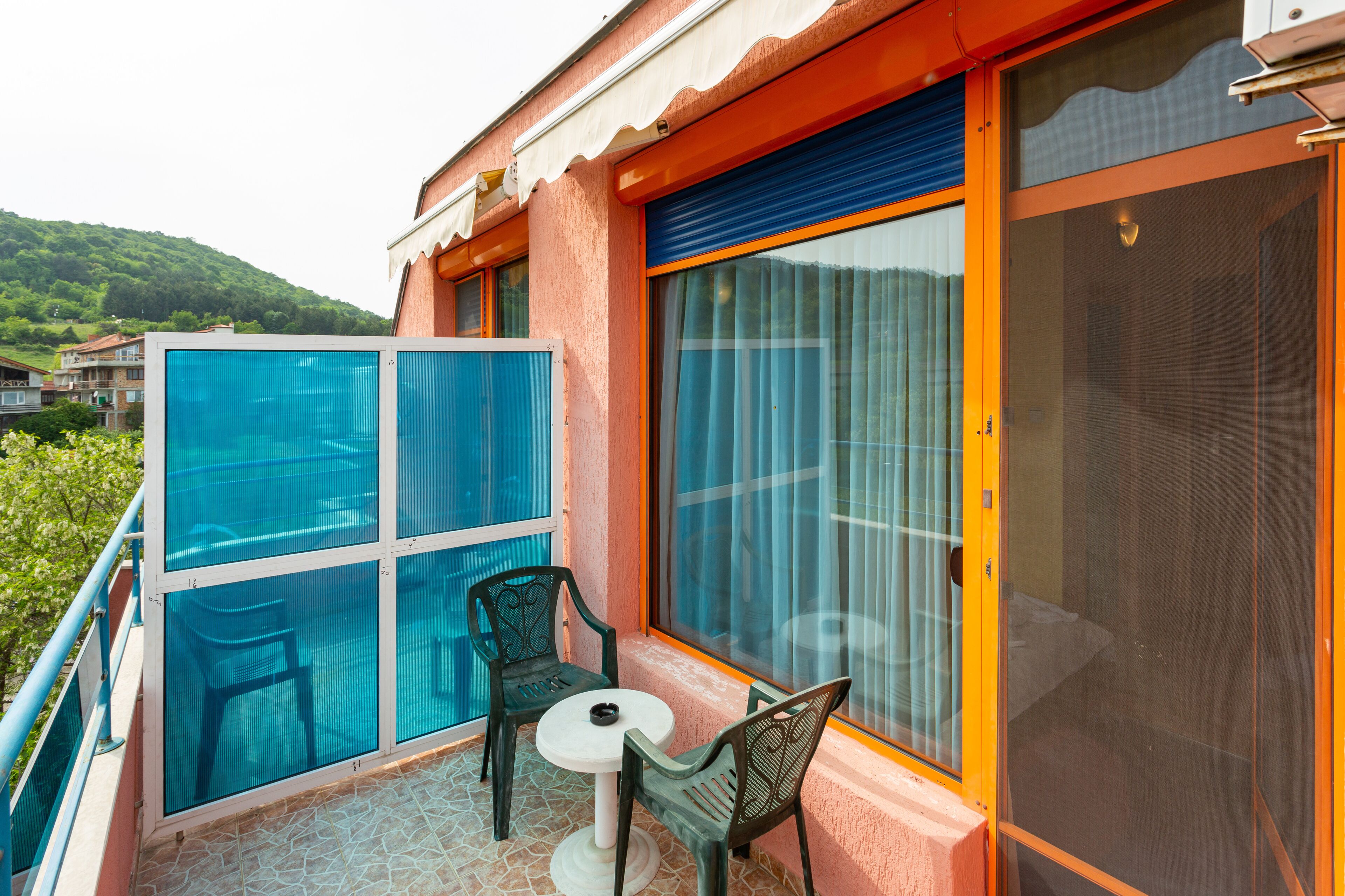 A balcony with two chairs and a table, a large window with curtains, and a view of a green hillside.