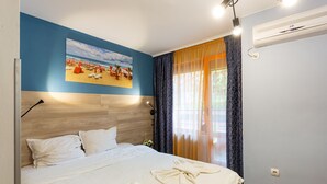 A hotel room with a bed, a painting of a beach scene, a window with curtains, and an air conditioning unit. - Family Hotel Jemelly (Obzor)
