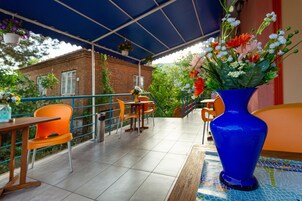 Un patio al aire libre con un paraguas azul, sillas anaranjadas y un florero azul con flores.
