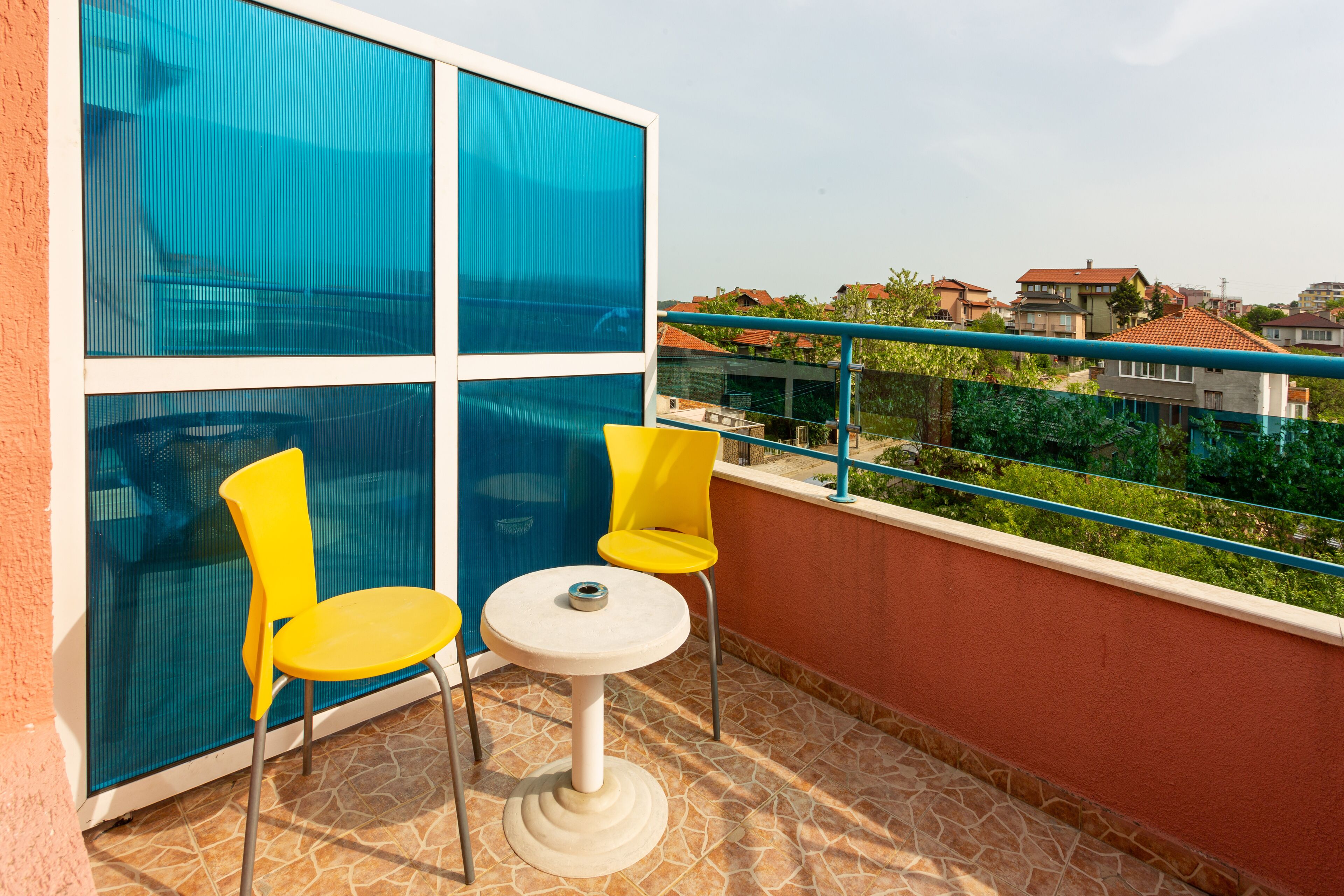 A balcony with two yellow chairs and a small table, overlooking a residential area with houses and greenery.