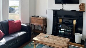 Una sala de estar con un sofá de cuero negro, una mesa de centro de madera, un televisor montado en la pared, una chimenea y un ventanal con vistas al exterior.