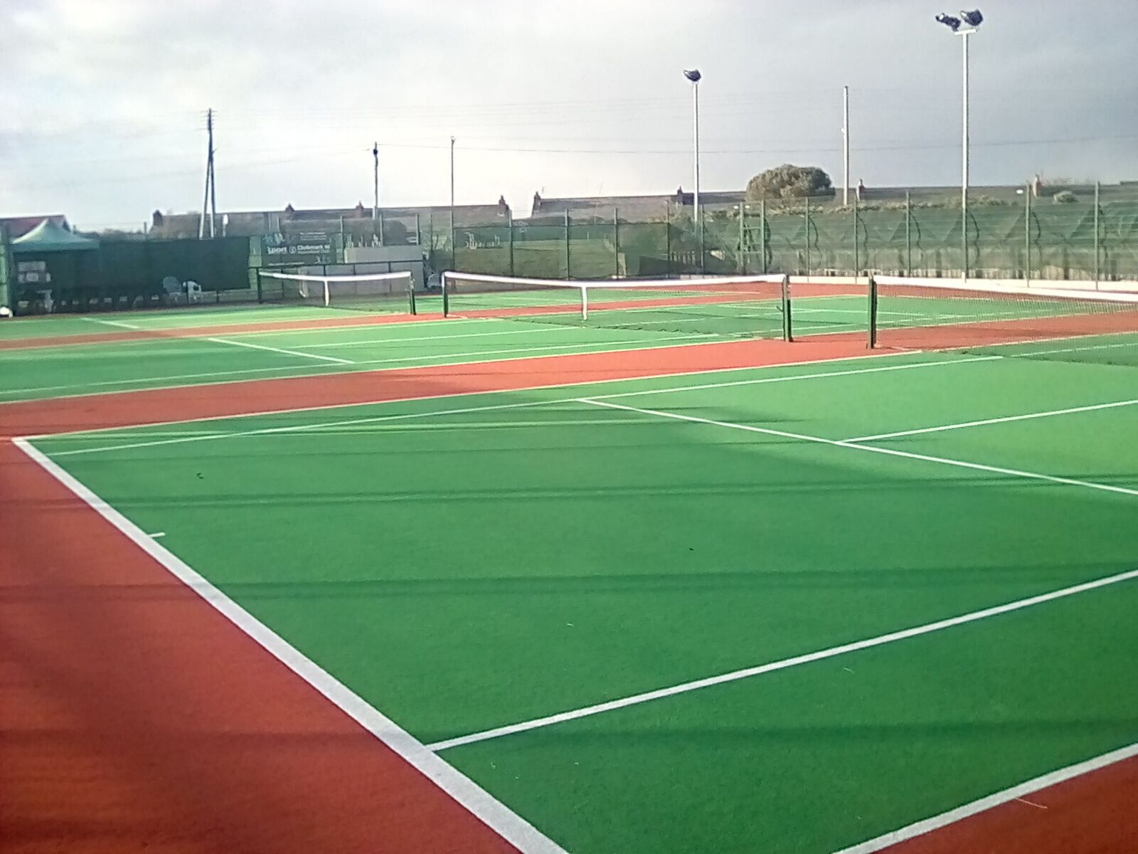 Een tennisbaan met meerdere groene en rode velden, omringd door hekken en lantaarns.