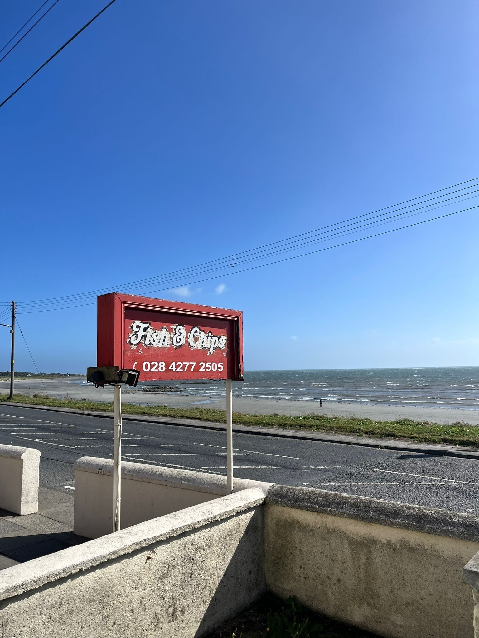 A red signboard for 'Fish & Chips' with a contact number, located beside a road with a view of the sea.