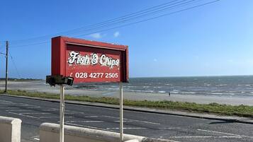 A red signboard for 'Fish & Chips' with a contact number, located beside a road with a view of the sea.