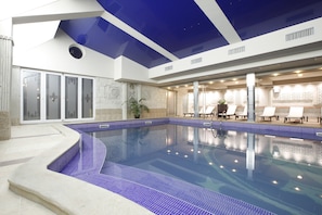 Una piscina interior con suelo y paredes de azulejos azules, techo transparente y sillones de descanso a lo largo de la piscina.